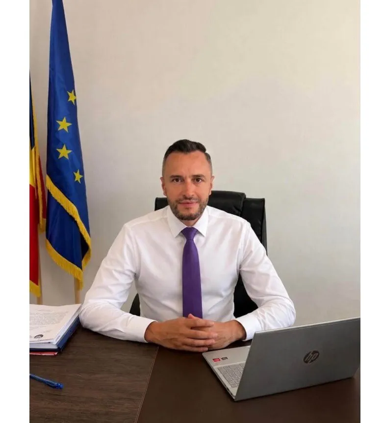 Un bărbat cu cămașă albă și cravată mov stă la un birou lângă un laptop și două steaguri.