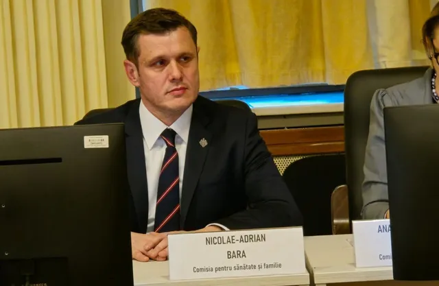 Un bărbat cu părul scurt, purtând un costum închis și o cravată în culorile albastru și roșu, stă la o masă cu un nume afișat.