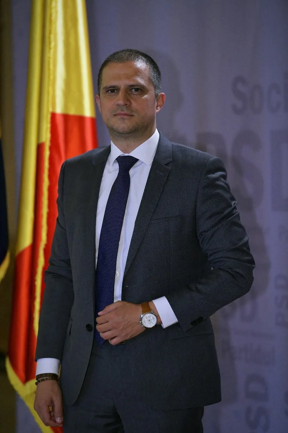 Un bărbat cu părul scurt poartă un costum gri, cămașă albă și cravată albastră, stând în fața unui steag galben și roșu.