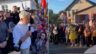 Un bărbat cu cămașă albă ține un microfon în fața unui grup de persoane strânse pe o stradă lângă clădiri.