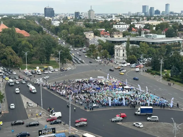 O mulțime de oameni este adunată pe o porțiune de asfalt într-o intersecție urbană mare, înconjurată de mașini, clădiri și copaci sub un cer înnorat.