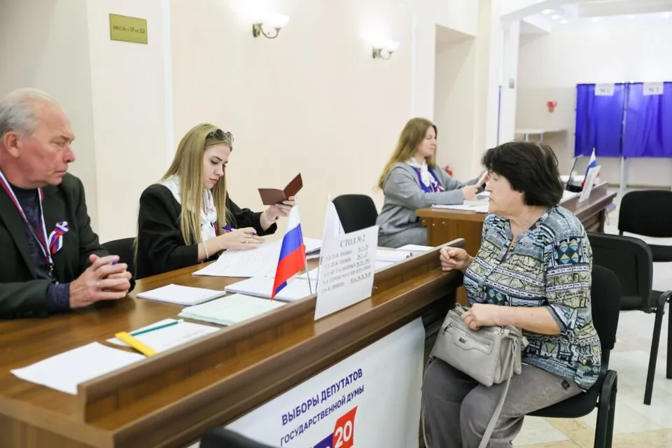 Trei persoane stau la mese de lemn în interiorul unei încăperi, în timp ce o femeie scrie pe o foaie de hârtie.