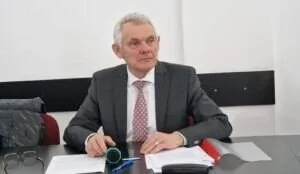 Un bărbat cu părul alb, purtând un sacou gri și o cravată roz, stă la o masă cu hârtii și ochelari în față.