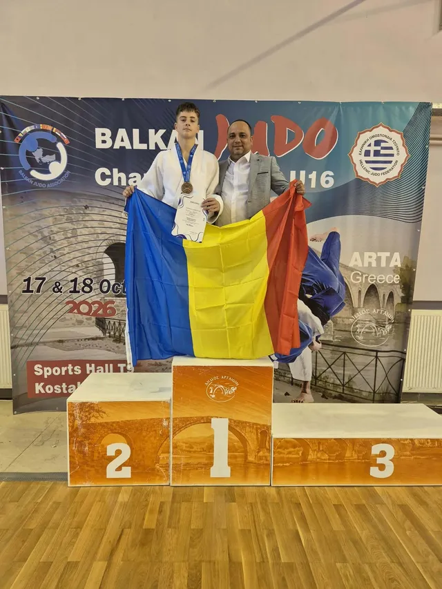 Un băiat cu o medalie la gât și un bărbat stau în spatele unui podium de premii colorat în fața unui panou publicitar.
