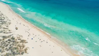 O vedere aeriană prezintă o plajă cu nisip deschis, unde persoane sunt împrăștiate pe mal și în apele turcoaz ale oceanului.