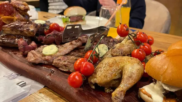 Un plat de carne gătită, cu roșii cherry și burgeri, este prezentat pe o tocătorie de lemn lângă un pahar cu suc portocaliu.