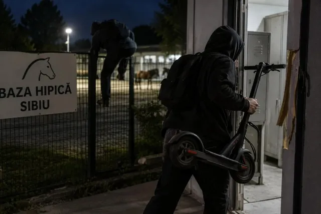 Un bărbat cu rucsac negru ține o trotinetă electrică în timp ce trece printr-o ușă deschisă, într-o scenă nocturnă.