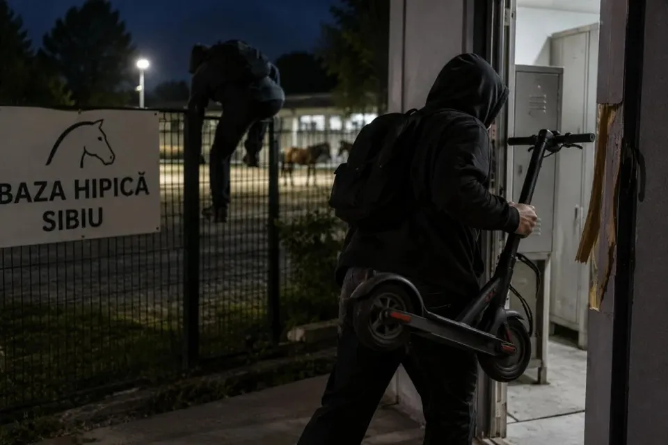 Un bărbat cu rucsac negru ține o trotinetă electrică în timp ce trece printr-o ușă deschisă, într-o scenă nocturnă.