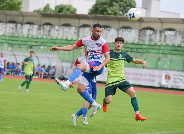 Un jucător de fotbal în echipament alb, roșu și albastru sare pentru a lovi o minge în aer pe un teren de fotbal.