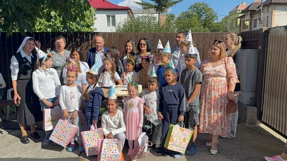Un grup de adulți și copii stau împreună în curte, mulți dintre copii ținând sacoșe colorate cu modele florale.