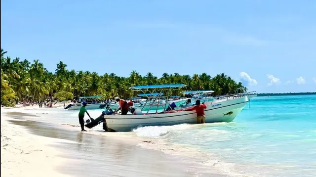 O barcă albă cu acoperiș albastru este trasă pe o plajă nisipoasă de către mai mulți oameni, lângă apele turcoaz ale mării.