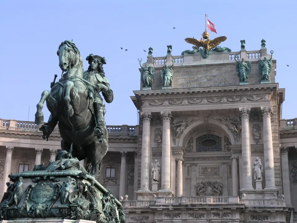O statuie ecustă a unui bărbat pe un cal se află în prim-plan, în fața unei clădiri monumentale cu coloane și sculpturi pe acoperiș.