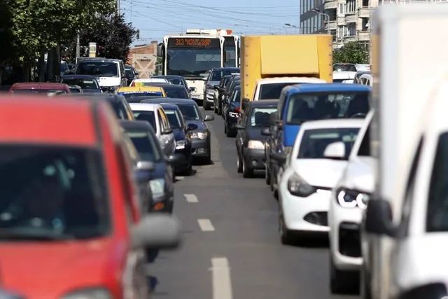 Mulți autovehicule, inclusiv camioane și mașini de pasager, sunt așezate în trafic pe un drum asfaltat cu linii de demarcare albe.