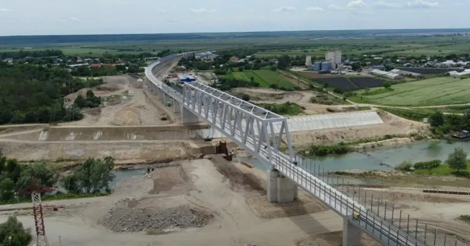 O structură metalică de tip pod traversează un curs de apă și un teren de șantier cu pământ și vegetație în fundal.