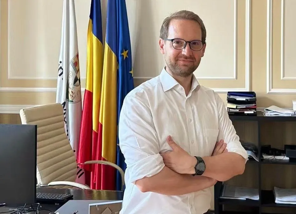 Un bărbat cu ochelari și cămașă albă stă cu brațele încrucișate în fața unor steaguri și al unui birou de birou.