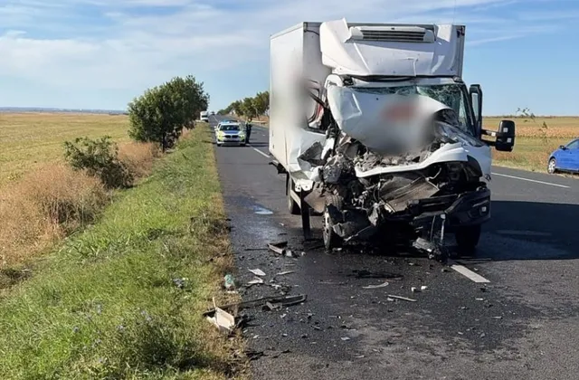 O autoutilitară albă este stopată pe marginea unui drum asfaltat, având partea frontală puternic zdrobită, în timp ce resturi sunt împrăștiate pe carosabil.