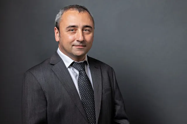 Un bărbat cu părul scurt și gri poartă un sacou gri închis, cămașă albă și o cravată neagră cu puncte mici, pe un fundal gri uniform.