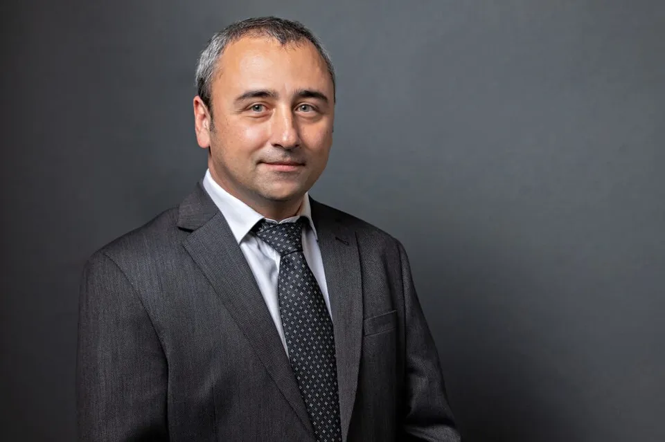 Un bărbat cu părul scurt și gri poartă un sacou gri închis, cămașă albă și o cravată neagră cu puncte mici, pe un fundal gri uniform.