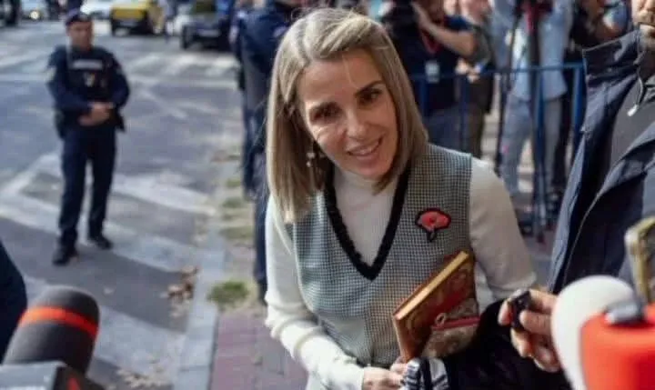 O femeie cu părul blond și vestă gri stă în picioare în fața unor microfoane, zâmbind ușor.