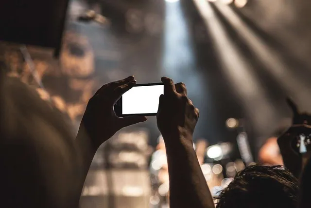 Două mâini țin un smartphone cu ecranul alb, capturând o scenă iluminată de proiectoare într-un spațiu întunecat.