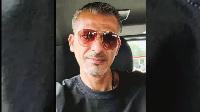 Un bărbat cu părul scurt și gri, purtând ochelari de soare cu lentile roz și un tricou negru, stă în interiorul unui autovehicul.