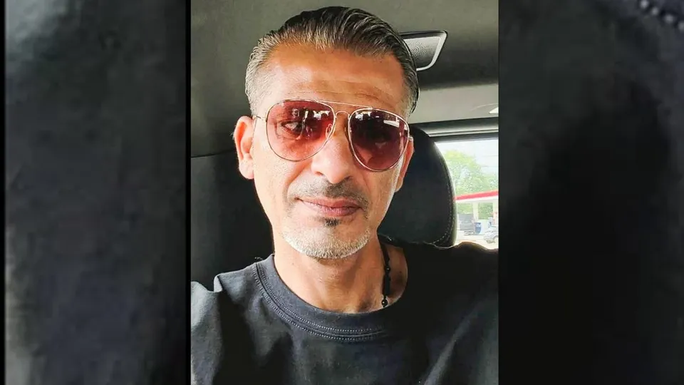 Un bărbat cu părul scurt și gri, purtând ochelari de soare cu lentile roz și un tricou negru, stă în interiorul unui autovehicul.