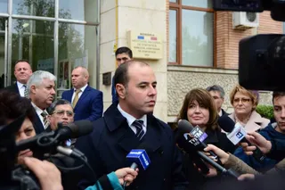Un bărbat cu părul scurt și haine închise răspunde unor microfoane de la un grup de persoane aflate în fața unei clădiri cu ferestre mari.