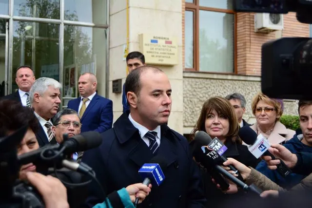 Un bărbat cu părul scurt și haine închise răspunde unor microfoane de la un grup de persoane aflate în fața unei clădiri cu ferestre mari.