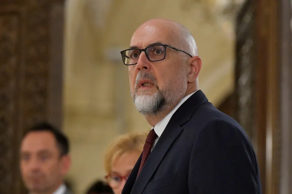Un bărbat cu ochelari, barbă scurtă și costum închis la culoare privește spre lateral într-un spațiu cu fundal neclar și decorات de lemn.