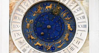 Un ceas circular cu fundal albastru prezintă diverse simboluri ale zodiacului și cifre romane pe un inel exterior alb.
