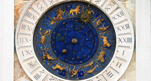 Un ceas circular cu fundal albastru prezintă diverse simboluri ale zodicilor aurii dispuse pe un cadran cu cifre romane pe margini albe.