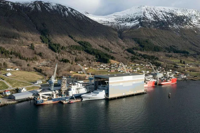 O vedere aeriană a unui șantier naval situat la malul apei, cu nave, macarale și un hangar mare în fața unui deal împădurit și înzăpezit.