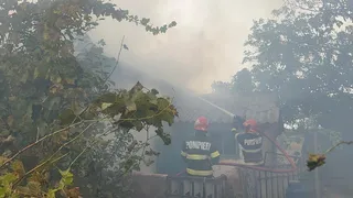 Doi pompieri cu echipament de protecție și cască roșie stau cu spatele, folosind un furtun pentru a stinge un incendiu la o clădire înzăpezită de fum.
