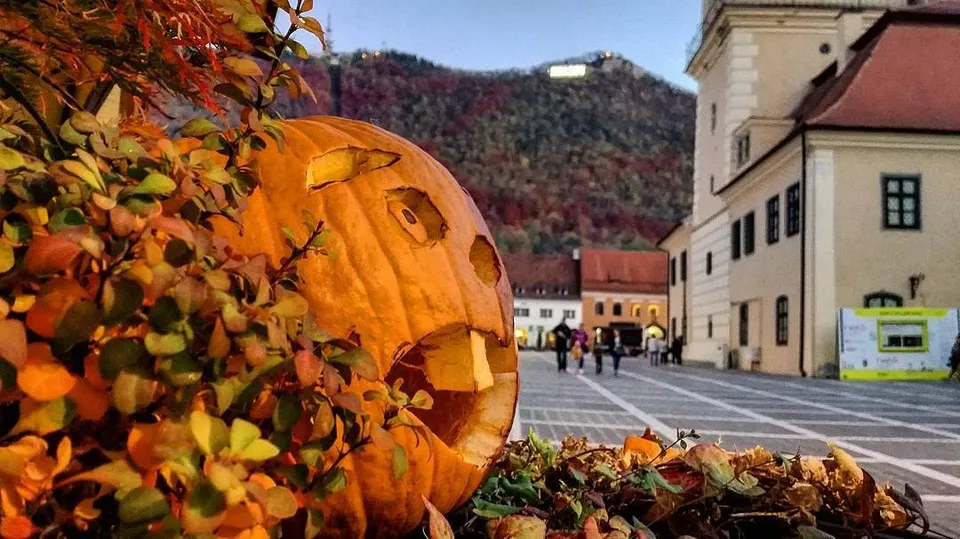 O pepenele de Halloween sculptată cu o față este situată lângă un tufiș de frunze colorate într-un oraș cu clădiri și oameni pe fundal.