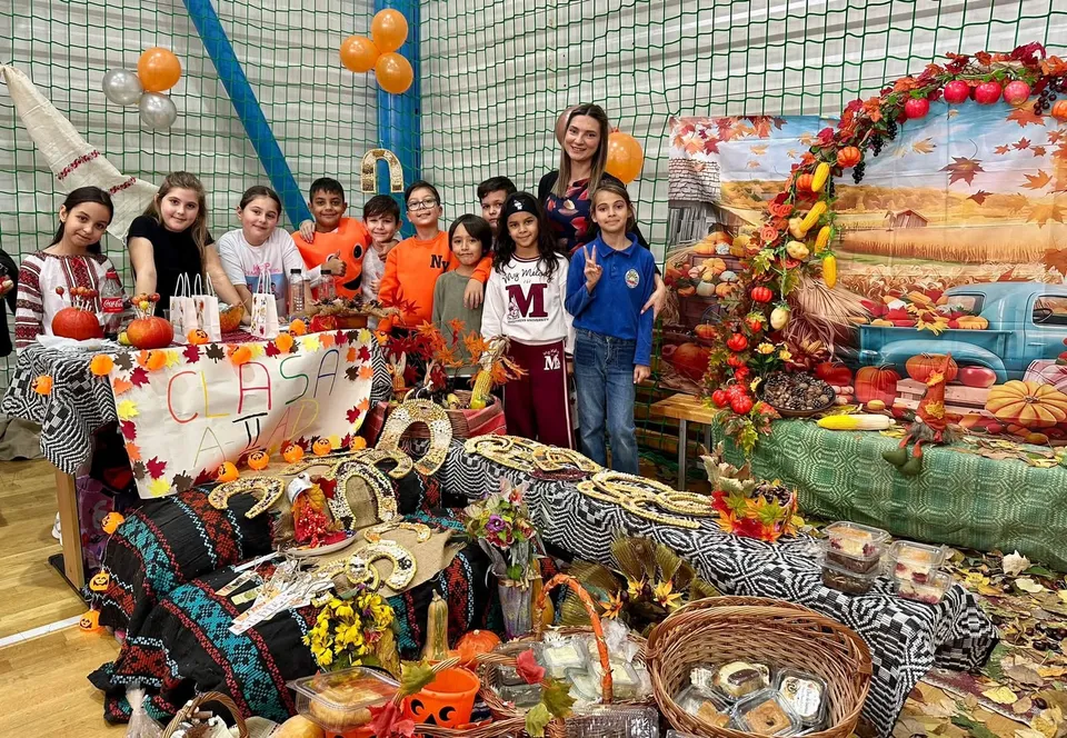 Un grup de copii și o femeie stau în spatele unor mese acoperite cu fructe, legume și obiecte decorative într-o încăpere cu plasă de protecție.