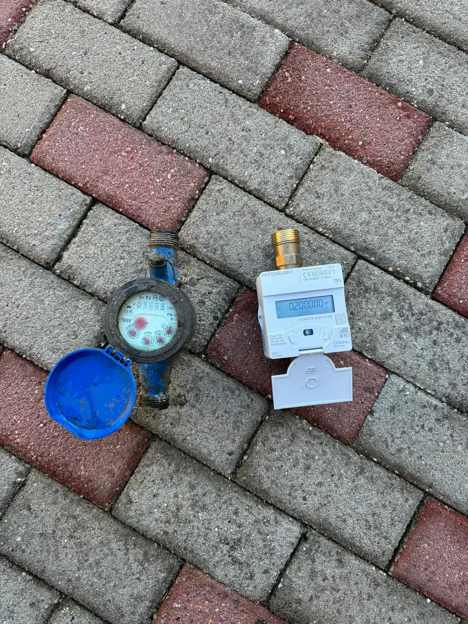 Un contor de apă digital alb și un contor mecanic cu capac albastru sunt așezate pe pavaj din cărămizi gri și roșii.