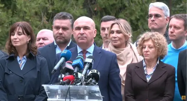 Un grup de persoane stă în spatele unui grup de microfoane dispuse pe un podium transparent, în fața unor copaci verzi.