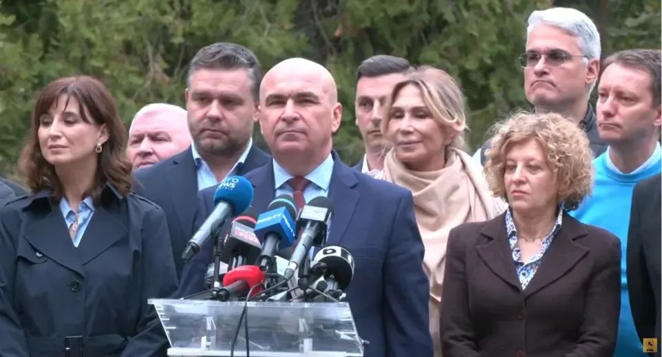 Un grup de persoane stă în spatele unui grup de microfoane dispuse pe un podium transparent, în fața unor copaci verzi.