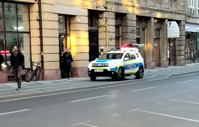 O mașină de poliție albă cu lumini de semnalizare aprinse stă pe un drum asfaltat lângă un trotuar și o clădire cu coloane.