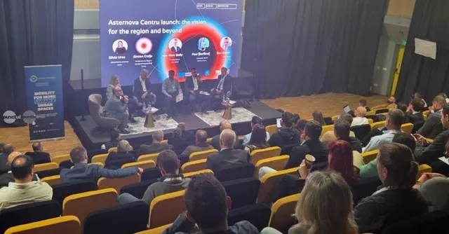 Oamenii stau așezați în scaune într-o sală de conferințe, privind spre un ecran mare pe care este afișat un grafic circular și patru persoane pe o scenă.