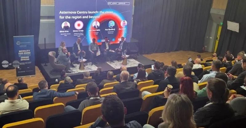 Oamenii stau așezați în scaune într-o sală de conferințe, privind spre un ecran mare pe care este afișat un grafic circular și patru persoane pe o scenă.