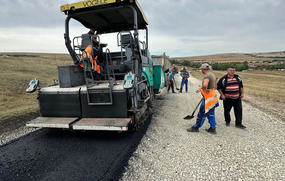 O mașină de asfalt este poziționată pe un drum de pietriș lângă un grup de lucrători care stau pe marginea drumului.