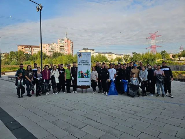 Un grup de oameni stă în rând pe o esplanadă pavată lângă un panou publicitar alb și vegetație.