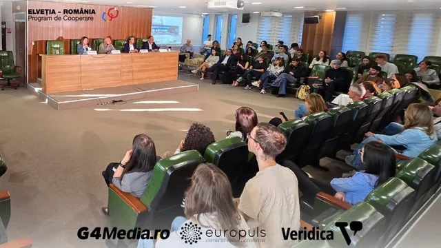 Oamenii stau așezați în scaunele unui auditorium, privind spre o masă lungă de lemn unde patru persoane sunt poziționate în partea din față.