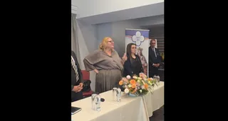 O femeie cu părul blond și o femeie cu părul închis stau în spatele unei mese acoperite cu un material alb, lângă un banner și flori.