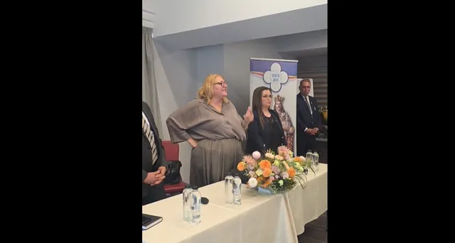 O femeie cu părul blond și o femeie cu părul închis stau în spatele unei mese acoperite cu un material alb, lângă un banner și flori.