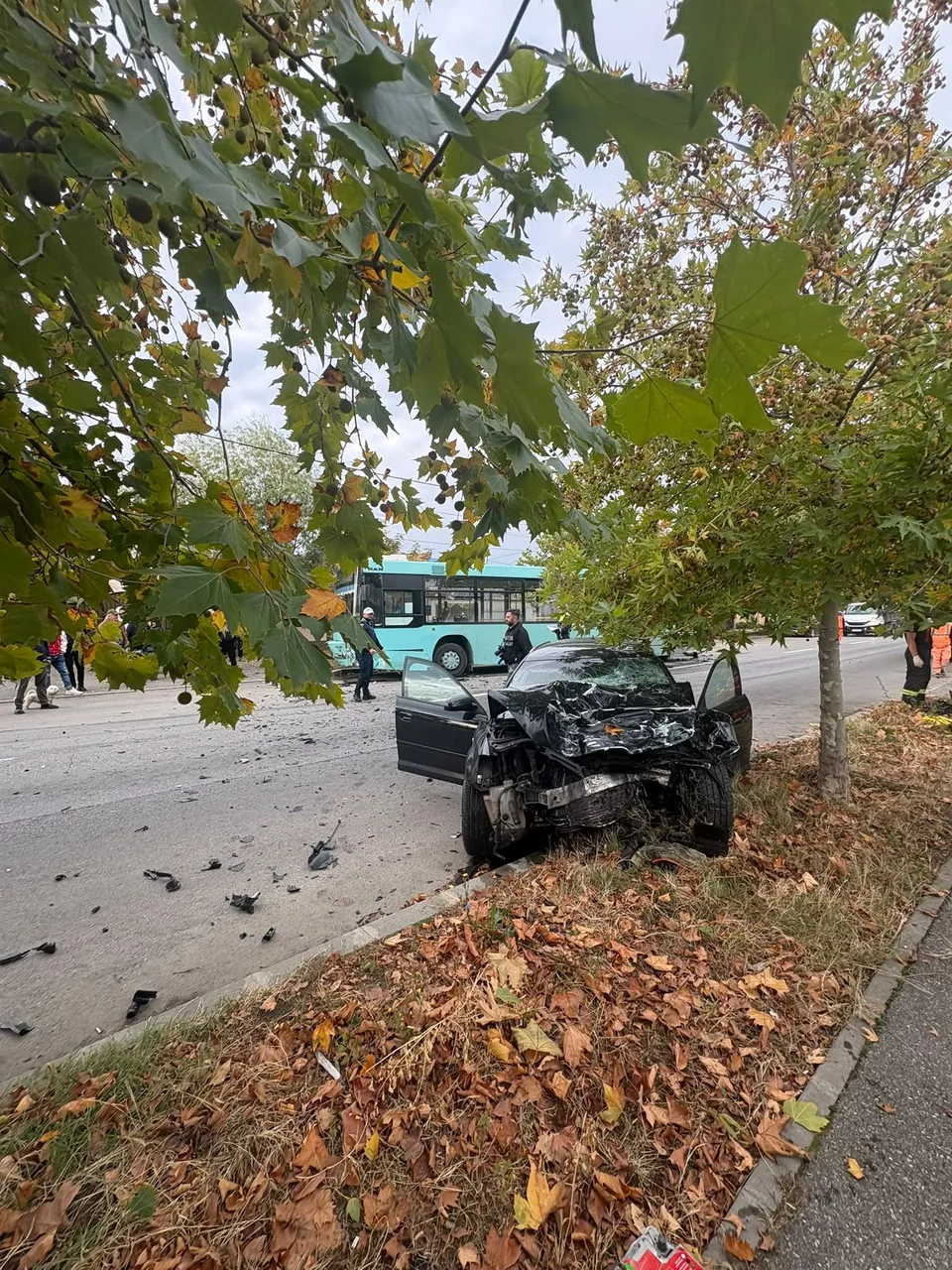 Un autoturism negru avariat stă pe marginea drumului lângă un autobuz turcoaz sub ramurile unor copaci cu frunze verzi și galbene.