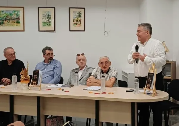Un bărbat într-o cămașă albă ține un microfon în timp ce cinci bărbați stau la o masă cu cărți și pahare de apă.