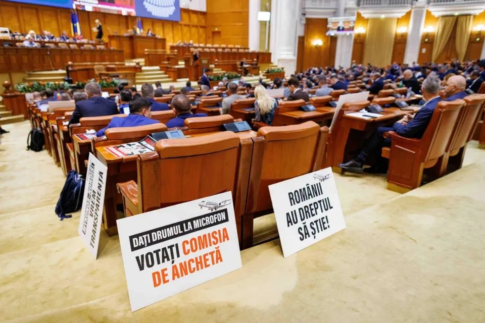 Câteva pliante albe cu text negru și roșu sunt așezate pe podeaua și pe scaunele din fața unor rânduri de scaune de lemn maro.