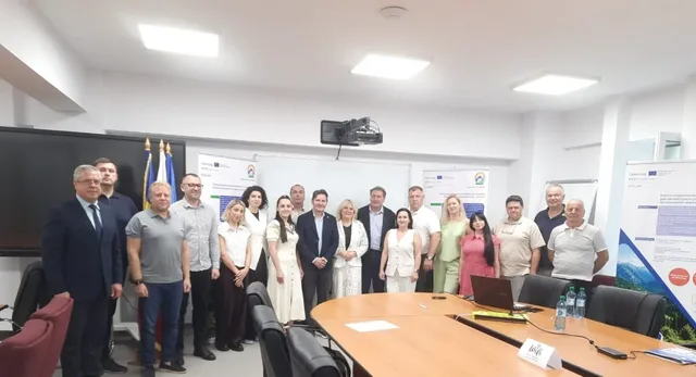 Un grup de aproximativ 20 de persoane, bărbați și femei, stau în picioare într-o sală de conferințe, în fața unei mese lungi. În fundal se văd steaguri și postere.